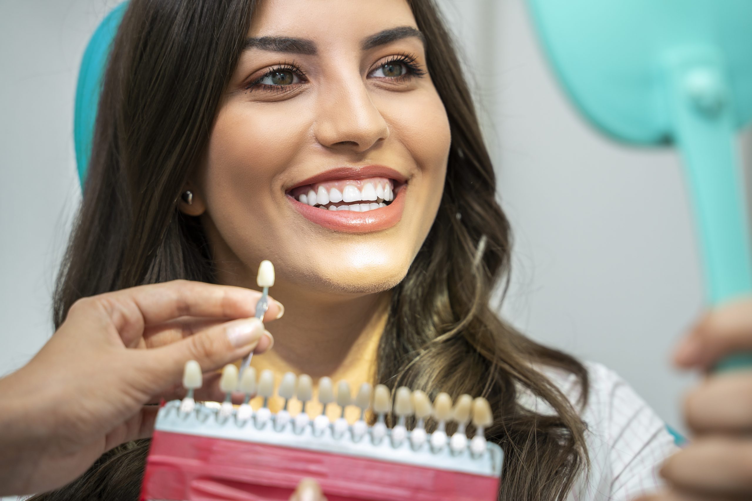 Smiling woman looking at her reflection in a dental chair while a dentist holds a false tooth up to show her the color comparison