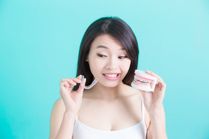 Woman looking at Invisalign aligner in her right hand with an indecisive look while holding fake teeth with braces in her left hand 