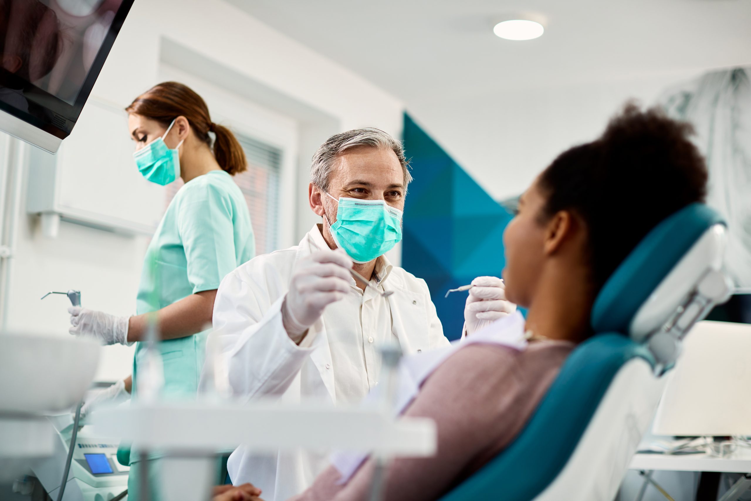 Dentist holding tools while looking at his patient and dental hygienist is in the background.