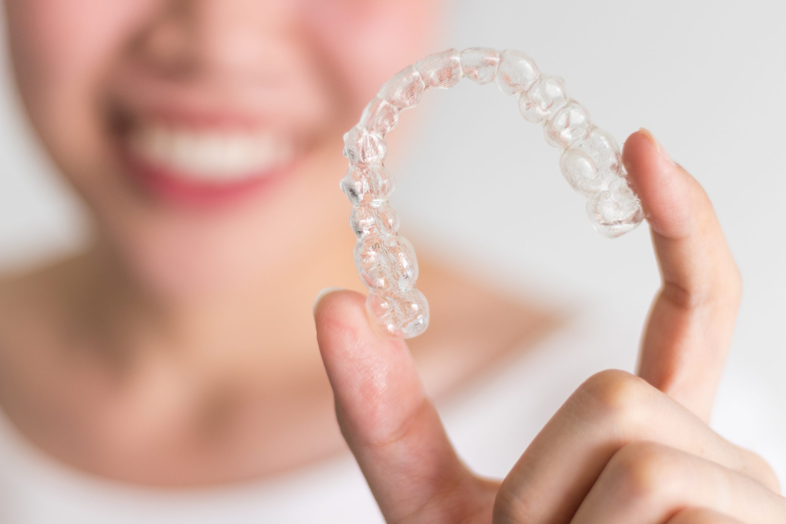 Smiling woman in a white shirt holding an Invisalign aligner between her pointer finger and thumb.