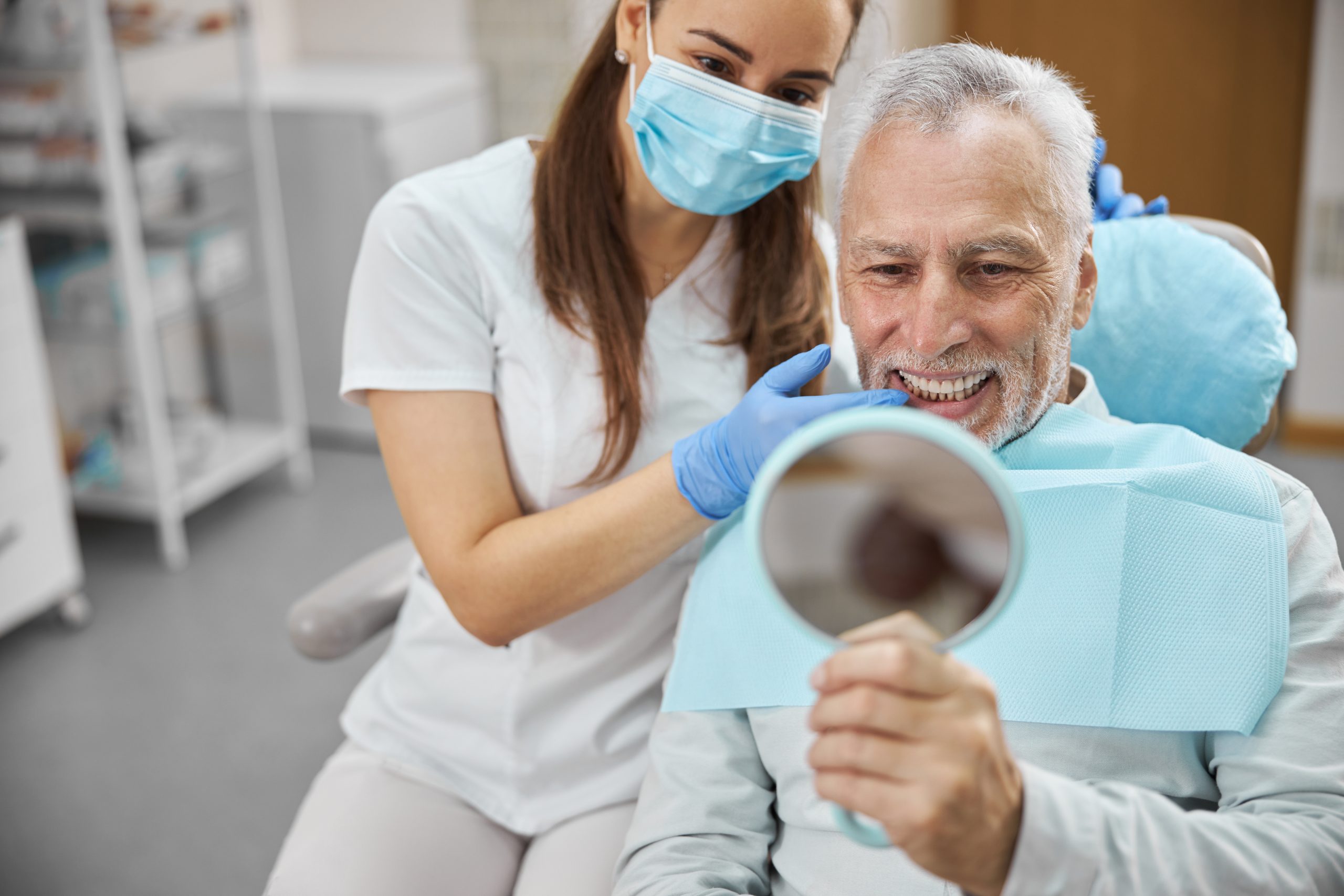 Patient admiring his new smile in a handheld mirror with a dentist behind him.