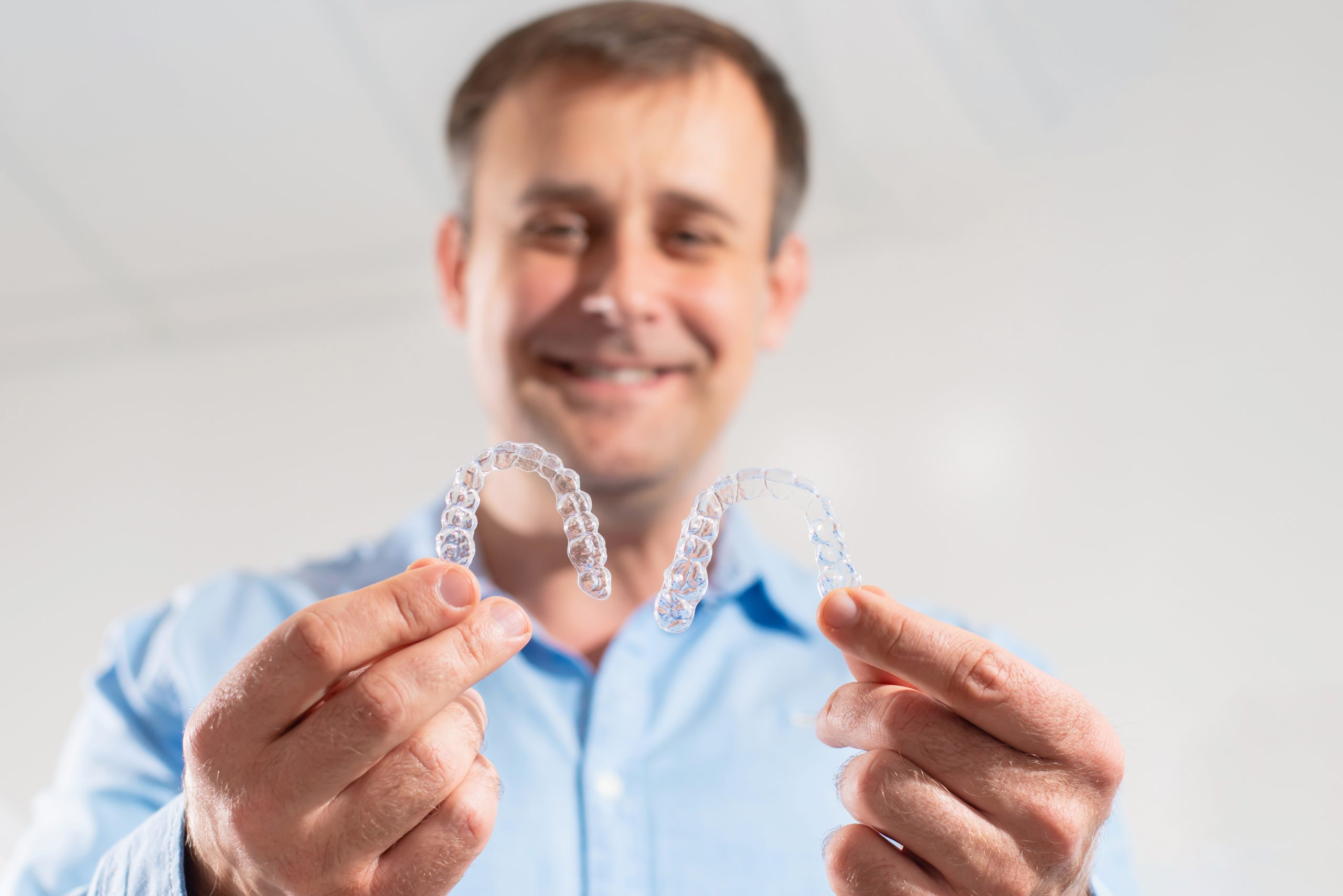 Smiling man in a blue button-down shirt holding an Invisalign aligner in each one of his hands with outstretched arms.
