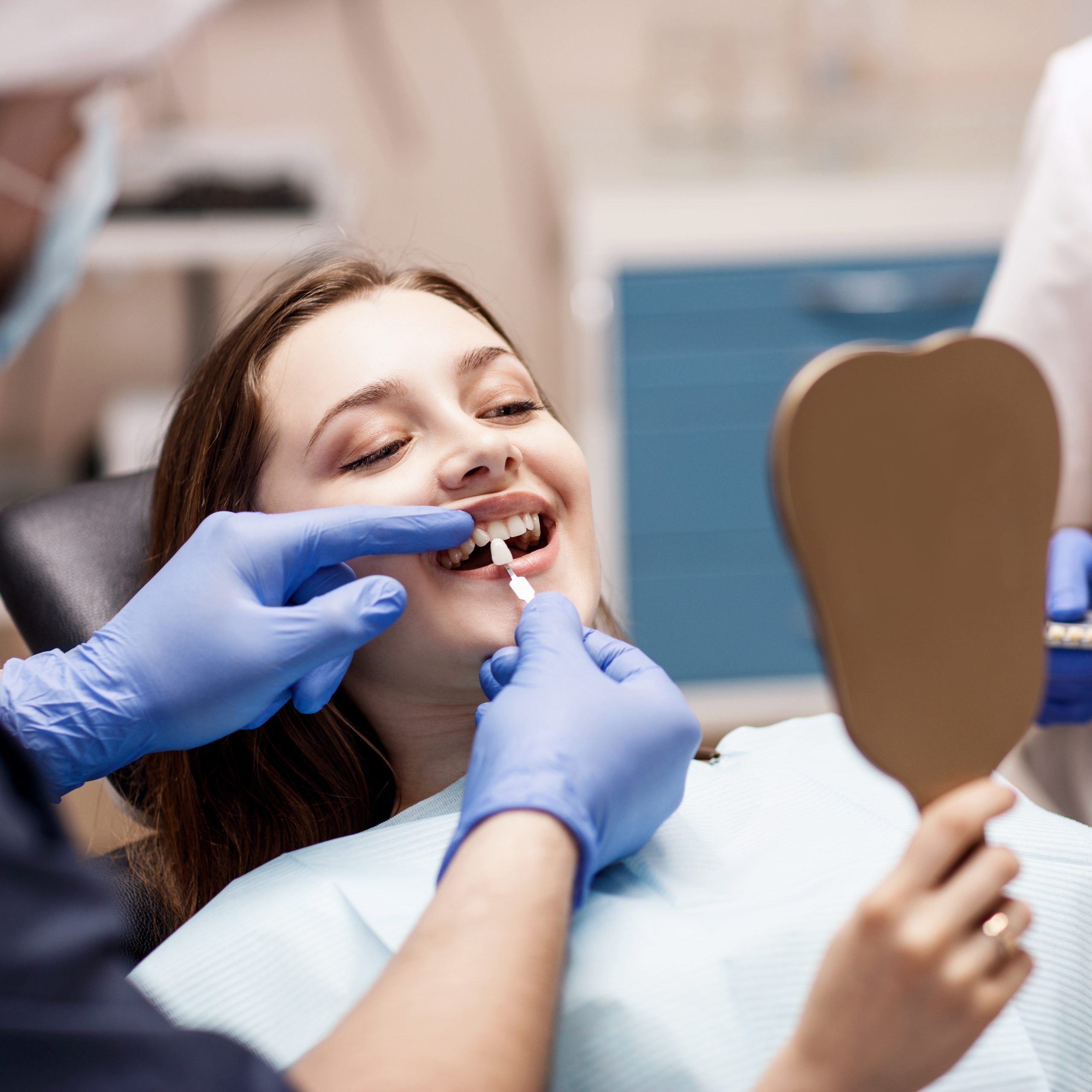 Woman looking in a dental chair looking at a handheld mirror while a dentist holds an artificial tooth to her mouth for color comparison.
